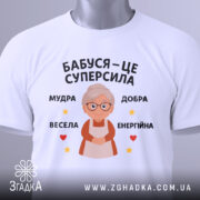 Футболка бабуся це суперсила, біла футболка, зображена зверху, на фоні світло-сірого кольору, принт з бабусею та текстом 'Бабуся – це суперсила', з зеленим ярликом, що звисає з коміра.