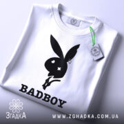 Футболка badboy кролик білого кольору, зображена зверху, на ній видно етикетку з зеленим шнурком, фон світло-сірий, принт чорного кольору з силуетом кролика та написом 'BADBOY'.