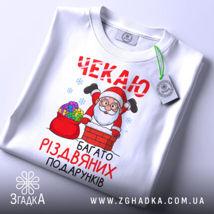 Футболка багато різдвяних подарунків, біла, складена на поверхні, фон світлий, принт з дідом Морозом, що виглядає з димаря, з кольоровими подарунками в мішку, з етикеткою.