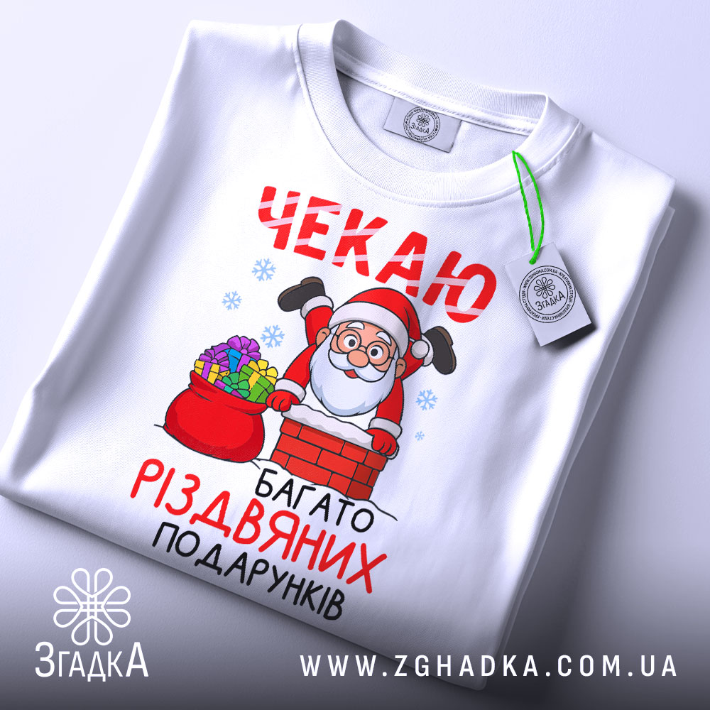 Футболка багато різдвяних подарунків, біла, складена на поверхні, фон світлий, принт з дідом Морозом, що виглядає з димаря, з кольоровими подарунками в мішку, з етикеткою.