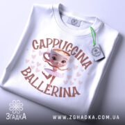 Футболка Ballerina Cappuccino білого кольору, складена на поверхні, з видимим етикеткою. Принт з малюнком балерини в центрі, оточено сердечками. Фон світло-сірий.