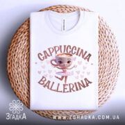 Футболка Ballerina Cappuccino білого кольору, складена на плетеній кошику. Принт з малюнком балерини в центрі, оточено сердечками. Фон світло-сірий.