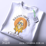 Футболка банан добрі руки, біла, зображена зверху, на столі, фон світло-сірий, принт з жовтим бананом у окулярах та написом 'Віддам свій банан в добрі руки....', з зеленим ярликом.