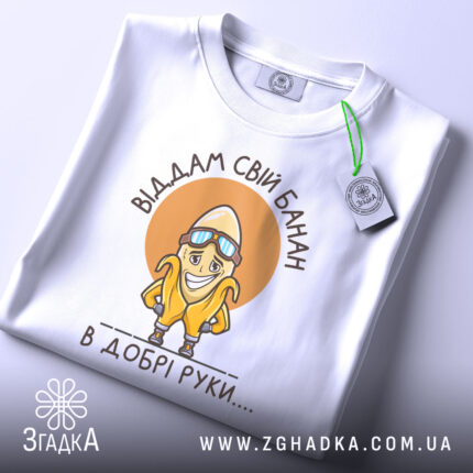 Футболка банан добрі руки, біла, зображена зверху, на столі, фон світло-сірий, принт з жовтим бананом у окулярах та написом 'Віддам свій банан в добрі руки....', з зеленим ярликом.
