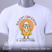 Футболка банан добрі руки, біла, зображена зверху, на столі, фон світло-сірий, принт з жовтим бананом у окулярах та написом 'Віддам свій банан в добрі руки....', з зеленим ярликом.