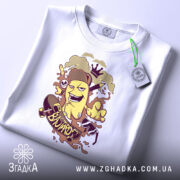 Футболка банан скейтер білого кольору, зображена зверху, на столі, фон світло-сірий, принт з жовтим бананом у шапці, що катається на скейтборді, з веселими елементами, на футболці є етикетка з зеленим шнурком.