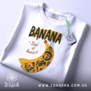 Футболка банан тату білого кольору, складена на столі, ракурс зверху, фон світло-сірий, принт з жовтою бананою та чорними написами, деталі принта включають малюнки черепів та інших елементів, на футболці є етикетка з зеленим шнурком.