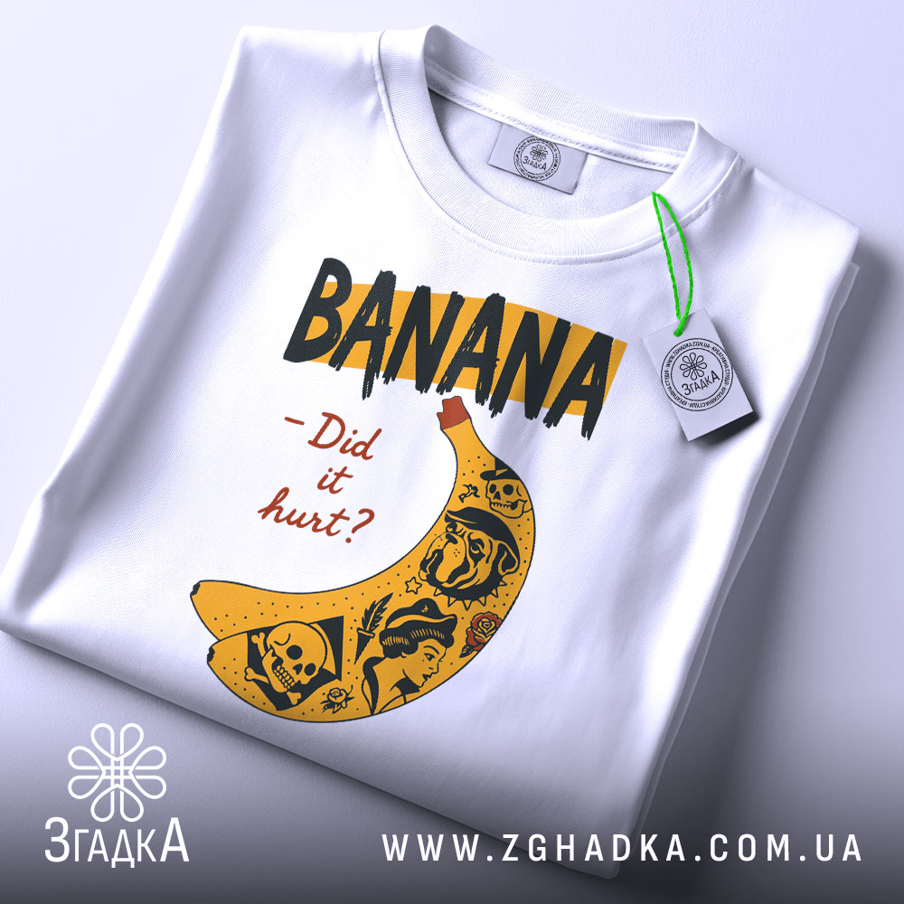 Футболка банан тату білого кольору, складена на столі, ракурс зверху, фон світло-сірий, принт з жовтою бананою та чорними написами, деталі принта включають малюнки черепів та інших елементів, на футболці є етикетка з зеленим шнурком.