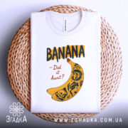 Футболка банан тату білого кольору, складена на плетеному кошику, ракурс зверху, фон світло-сірий, принт з жовтою бананою та чорними написами, деталі принта включають малюнки черепів та інших елементів.
