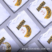 Футболка банан тату білого кольору, зображена на манекені, ракурс спереду, фон світло-сірий, принт з жовтою бананою та чорними написами, деталі принта включають малюнки черепів та інших елементів.