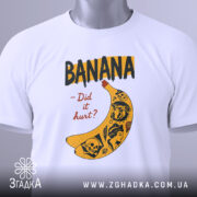 Футболка банан тату білого кольору, складена на столі, ракурс зверху, фон світло-сірий, принт з жовтою бананою та чорними написами, деталі принта включають малюнки черепів та інших елементів, на футболці є етикетка з зеленим шнурком.