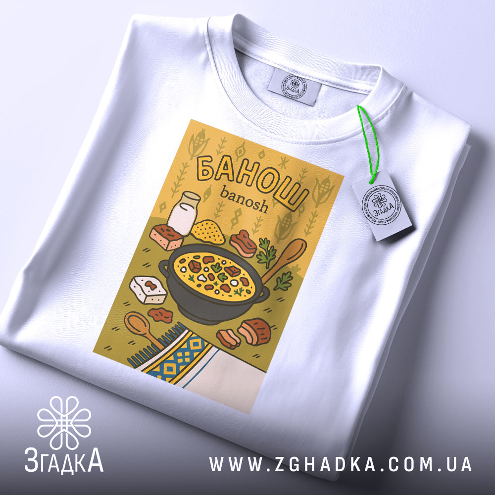 Футболка банош українська кухня, біла, складена на столі, фон світло-сірий, принт з жовтим фоном, на якому зображена каструля з баношем, молоко, хліб та інші інгредієнти, з зеленим ярликом.