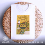 Футболка банош українська кухня, біла, складена на плетеному кошику, фон світло-сірий, принт з жовтим фоном, на якому зображена каструля з баношем, молоко, хліб та інші інгредієнти.