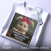 Футболка бантик тримає спокій, біла, складена на столі, фон світлий, принт з кроликом з рожевим бантиком і текстом на чорному фоні, з етикеткою на рукаві.