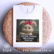 Футболка бантик тримає спокій, біла, складена на кошику, фон натуральний, принт з кроликом з рожевим бантиком і текстом на чорному фоні.