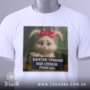 Футболка бантик тримає спокій, біла, складена на столі, фон світлий, принт з кроликом з рожевим бантиком і текстом на чорному фоні, з етикеткою на рукаві.