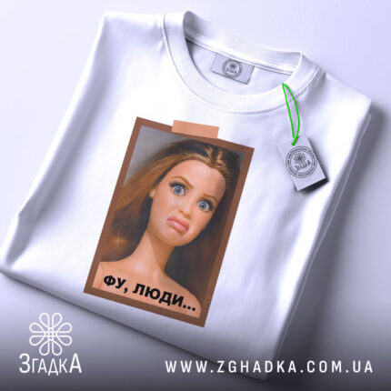 Футболка барбі фу люди. Біла футболка, складена на столі, з принтом ляльки, яка має вираз обличчя, що показує незадоволення. Принт оточений рамкою коричневого кольору, а на футболці є етикетка з зеленим шнурком. Фон світло-сірий.
