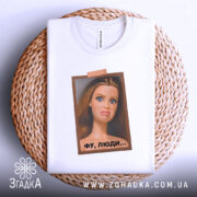 Футболка барбі фу люди. Біла футболка, складена на плетеній основі, з принтом ляльки, яка має вираз обличчя, що показує незадоволення. Принт оточений рамкою коричневого кольору, фон світло-сірий.