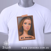 Футболка барбі фу люди. Біла футболка, складена на столі, з принтом ляльки, яка має вираз обличчя, що показує незадоволення. Принт оточений рамкою коричневого кольору, а на футболці є етикетка з зеленим шнурком. Фон світло-сірий.