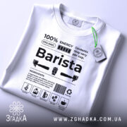 Футболка бариста стиль, біла, складена на столі, фон світло-сірий, принт чорного кольору з текстом та іконками, на футболці є етикетка з зеленим шнурком.