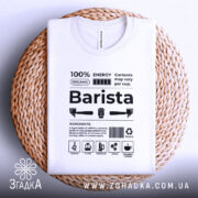 Футболка бариста стиль, біла, складена на плетеному кошику, фон світло-сірий, принт чорного кольору з текстом та іконками, що описують інгредієнти для приготування кави.