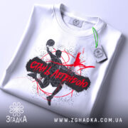 Футболка баскетбол стань легендою, біла футболка складена на поверхні, принт з баскетболістом у чорному та червоному кольорах, етикетка з зеленим шнурком, фон світлий, акцент на принті.