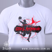 Футболка баскетбол стань легендою, біла футболка складена на поверхні, принт з баскетболістом у чорному та червоному кольорах, етикетка з зеленим шнурком, фон світлий, акцент на принті.