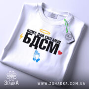 Футболка БДСМ білого кольору, зображена на столі, фон світло-сірий, принт містить текст "Боже дай сил мені БДСМ" з жовтим німбом, блискавкою, блакитними руками та червоним сердечком, на футболці є етикетка.