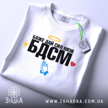 Футболка БДСМ білого кольору, зображена на столі, фон світло-сірий, принт містить текст "Боже дай сил мені БДСМ" з жовтим німбом, блискавкою, блакитними руками та червоним сердечком, на футболці є етикетка.