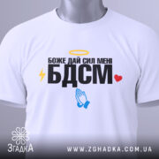 Футболка БДСМ білого кольору, зображена на столі, фон світло-сірий, принт містить текст "Боже дай сил мені БДСМ" з жовтим німбом, блискавкою, блакитними руками та червоним сердечком, на футболці є етикетка.