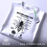 Футболка бджола принт білого кольору, складена на поверхні, фон світло-сірий, принт чорного кольору з зображенням бджоли та текстом, на футболці зелений ярлик.
