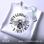Футболка бджоли і мед, білого кольору, складена на плоскій поверхні. Принт з бджолою та написом в чорному кольорі, з зеленим ярликом, фон світло-сірий.