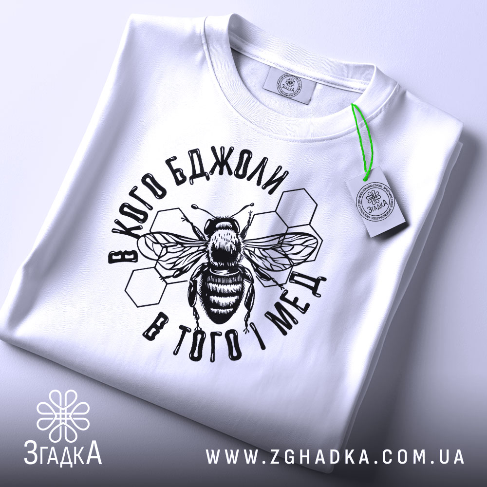 Футболка бджоли і мед, білого кольору, складена на плоскій поверхні. Принт з бджолою та написом в чорному кольорі, з зеленим ярликом, фон світло-сірий.