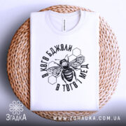 Футболка бджоли і мед, білого кольору, складена на плоскій поверхні з плетеного матеріалу. Принт з бджолою та написом в чорному кольорі, фон світло-сірий.