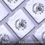 Футболка бджоли і мед, білого кольору, зображена на манекені з блискучою поверхнею, фон світло-сірий. Принт з бджолою та написом в чорному кольорі, оточений шестиугольниками.