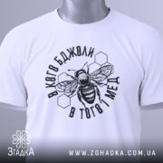 Футболка бджоли і мед, білого кольору, складена на плоскій поверхні. Принт з бджолою та написом в чорному кольорі, з зеленим ярликом, фон світло-сірий.