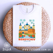 Футболка Бердичів, біла, зображена зверху на плетеному кошику, фон світло-сірий, принт з архітектурними елементами та квітами, текст 'BERDYCHIV UKRAINE' яскраво-оранжевий.