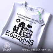 Футболка Бердичів надихає. Біла футболка, складена на поверхні, з видимим етикеткою. Принт на футболці містить текст та малюнки, фон світлий і чистий.