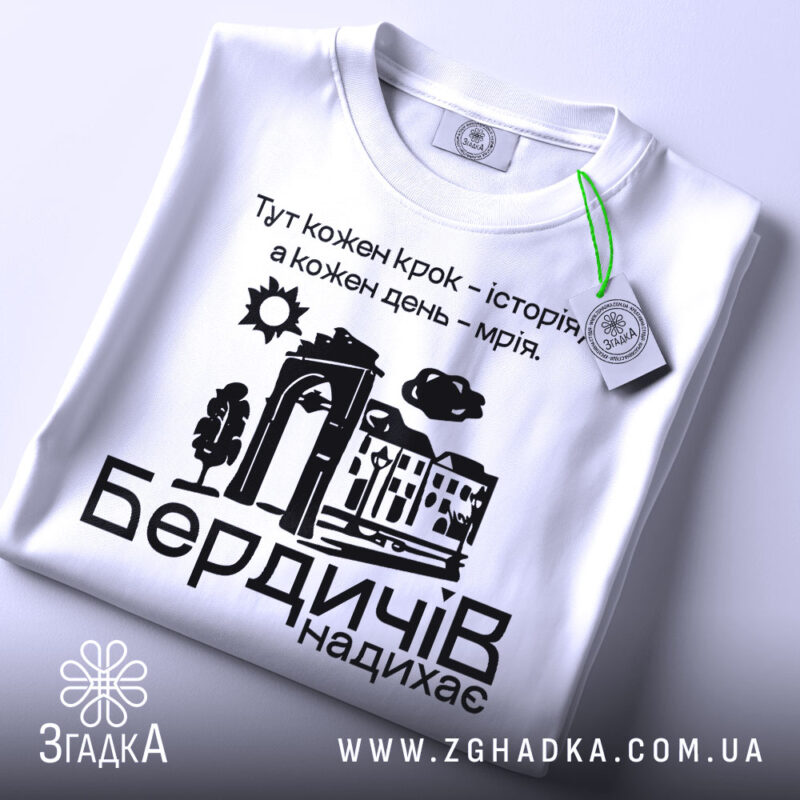 Футболка Бердичів надихає. Біла футболка, складена на поверхні, з видимим етикеткою. Принт на футболці містить текст та малюнки, фон світлий і чистий.