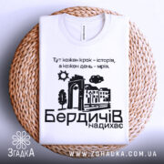 Футболка Бердичів надихає. Біла футболка, складена на плетеній кошику, фон білий. Принт містить текст і малюнки, що відображають архітектурні елементи та природу.