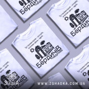 Футболка Бердичів надихає. Біла футболка з коротким рукавом, зображена на манекені, фон нейтральний. Принт містить текст і графічні елементи, що зображують архітектурні деталі та природу.