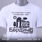 Футболка Бердичів надихає. Біла футболка, складена на поверхні, з видимим етикеткою. Принт на футболці містить текст та малюнки, фон світлий і чистий.