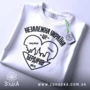 Футболка Бердичів подарунок, біла, складена на поверхні, фон світло-сірий, принт чорного кольору з написом 'Незалежна Україна - це Бердичів' та силуетом міста, з зеленим ярликом.
