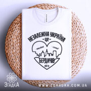 Футболка Бердичів подарунок, біла, складена на плетеній кошику, фон світло-сірий, принт чорного кольору з написом 'Незалежна Україна - це Бердичів' та силуетом міста.