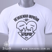 Футболка Бердичів подарунок, біла, складена на поверхні, фон світло-сірий, принт чорного кольору з написом 'Незалежна Україна - це Бердичів' та силуетом міста, з зеленим ярликом.