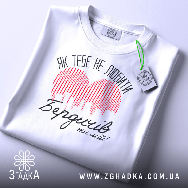 Футболка Бердичів білого кольору, складена на поверхні, з видимим етикеткою. Принт червоного кольору у формі сердечка з малюнком міста та текстом "Як тебе не любити Бердичів ти мій!" в чорному кольорі, фон світло-сірий.