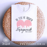 Футболка Бердичів білого кольору, складена на плетеній кошику, з видимим принтом червоного кольору у формі сердечка з малюнком міста та текстом "Як тебе не любити Бердичів ти мій!" в чорному кольорі, фон світло-сірий.