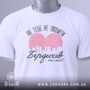 Футболка Бердичів білого кольору, складена на поверхні, з видимим етикеткою. Принт червоного кольору у формі сердечка з малюнком міста та текстом "Як тебе не любити Бердичів ти мій!" в чорному кольорі, фон світло-сірий.