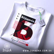 Футболка Бердичів заснований, біла, складена на поверхні, фон світлий, принт з червоним та чорним текстом, прикріплена етикетка з зеленим шнурком.