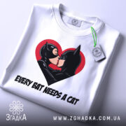 Футболка бетмен кішка кохання, біла, складена на столі, фон світло-сірий, принт з червоним серцем, на якому поцілуються персонажі Бетмен і Кішка, з етикеткою зеленого кольору.